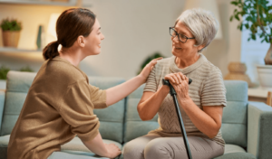 A young woman is seated, smiling and touching the shoulder of an older woman with glasses, also seated and holding a cane. Both are inside a well-lit room with plants and shelves in the background, showcasing the benefits of memory care in Memory Care Facilities.