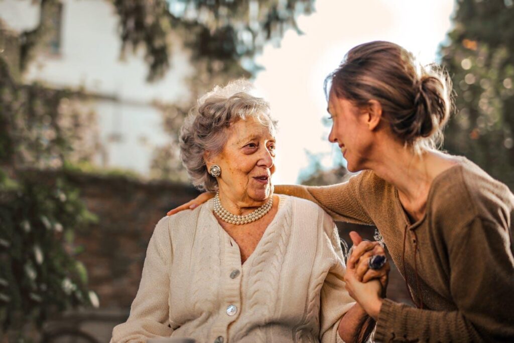 In the serene setting of Ocala, FL, an older woman and a younger woman sit outdoors, smiling warmly at each other. The older woman wears a cardigan and pearls while the younger gently holds her hand—a heartwarming moment in their assisted living community.