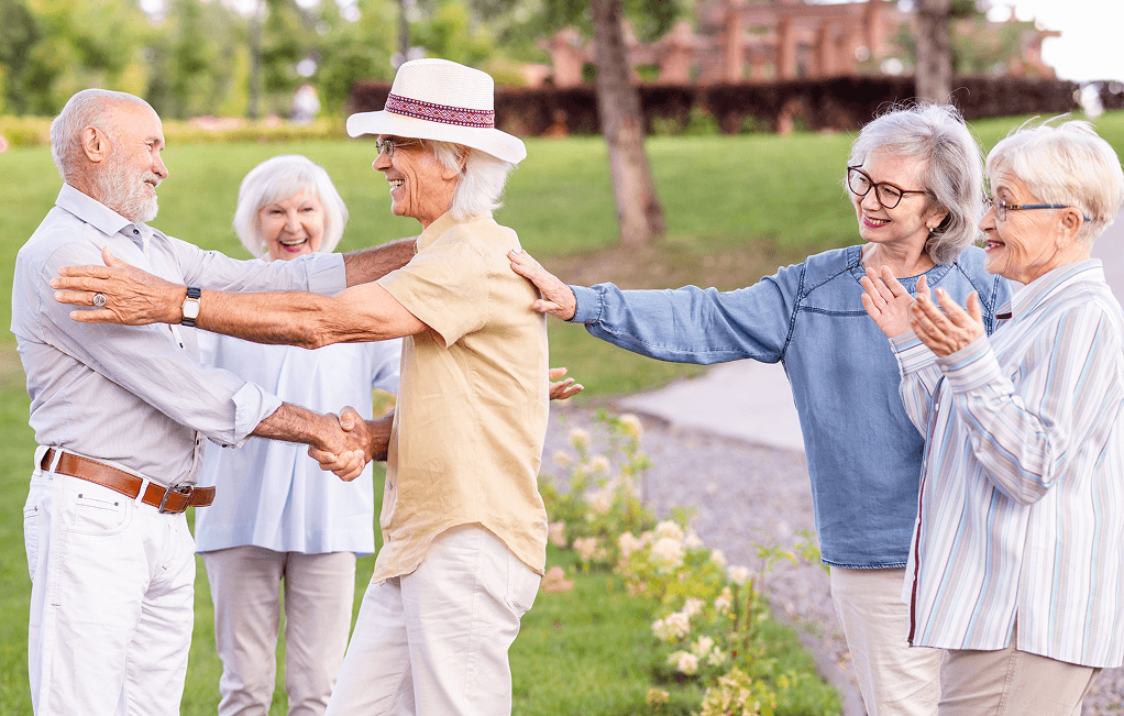 A group of five older adults is outdoors in Ocala, greeting each other with smiles and handshakes. They appear happy, embracing the warmth in their casual summer clothing. If you're seeking a similar lifestyle, consider exploring assisted living options tailored just for you.