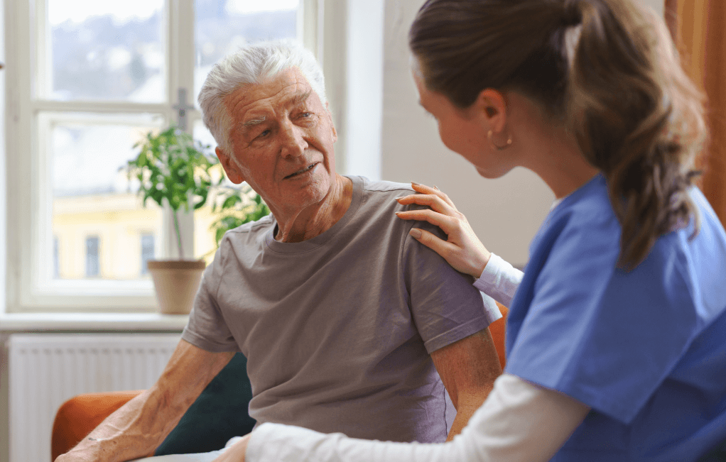 An elderly man in a grey shirt sits indoors, engaging with a woman in blue scrubs who is touching his shoulder supportively. This warm exchange reflects Florida's commitment to high-quality senior care.