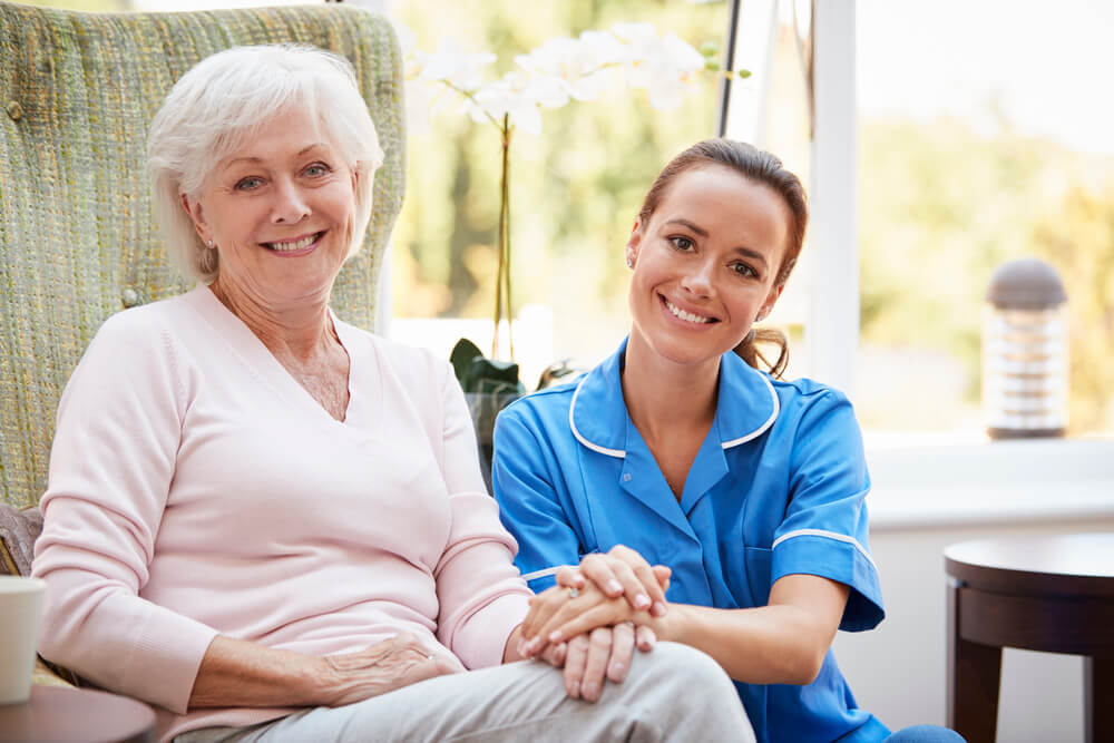 An older woman sits and smiles next to a nurse in blue scrubs at Hampton Manor assisted living. The nurse is holding the woman's hand, and both are looking at the camera, creating a warm atmosphere that feels like home.