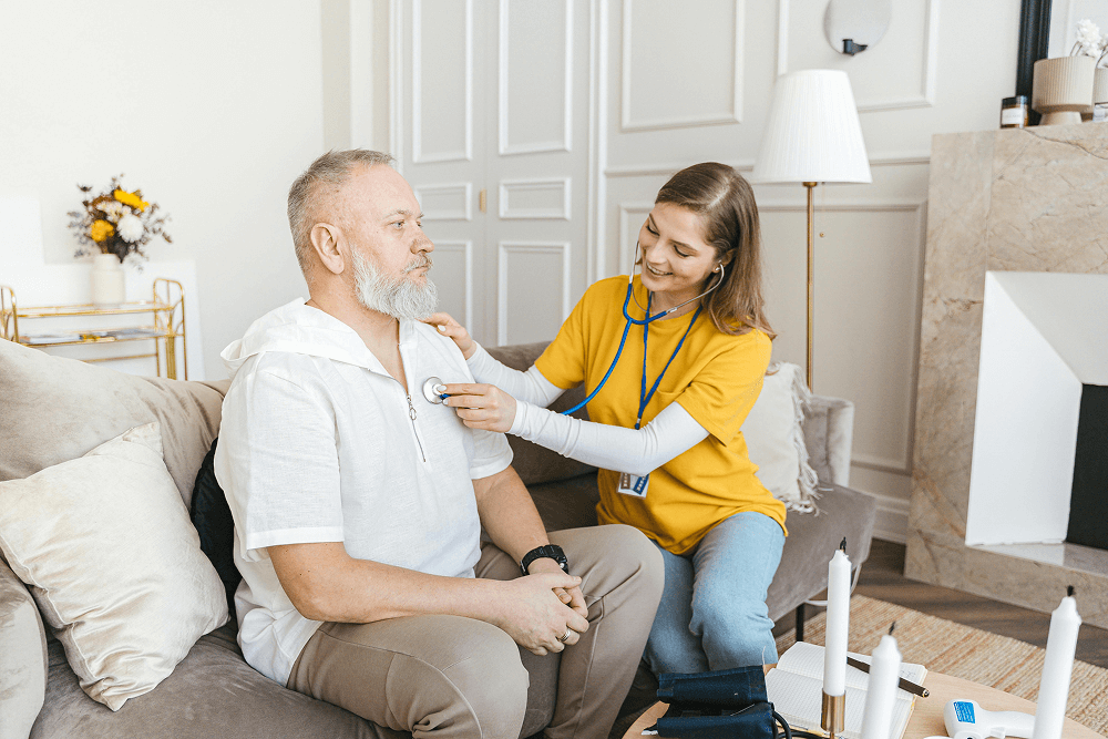 A healthcare worker uses a stethoscope to check the chest of an older man sitting on a couch, providing comfort and support as he transitions to memory care in a familiar living room setting.