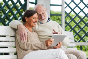 An older couple sits on a white bench outdoors, smiling and looking at a tablet together as they research how to pay for memory care in Florida.