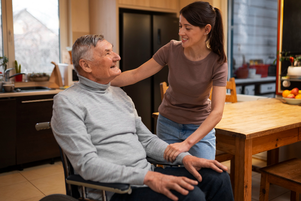 A young woman stands beside an older man in a wheelchair, smiling and holding his hand in a warm kitchen—capturing the caring spirit found in senior living Ocala.